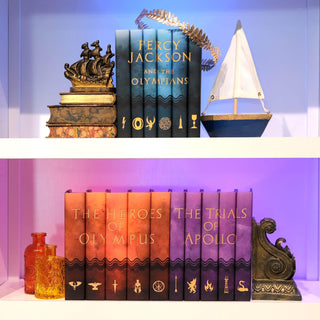 Percy Jackson and the Olympians book set on a white shelf sitting above the Heroes of Olympus, and The Trials of Apollo book sets surrounded by antique books, a sailboat, and and glass jars.
