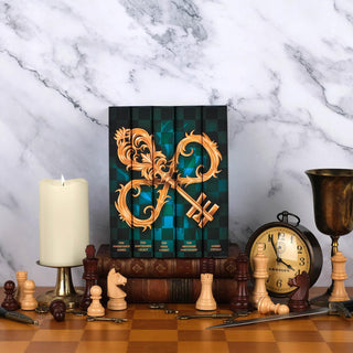 The Inheritance Games Series by Jennifer Lynn Barnes from Juniper Book sits on a stack of antique leather books sitting on a wooden chess board set against a white marble background. The stack is surrounded by chess pieces, a candle, an antique clock, a gold cup, a dagger, and ornate gold keys.