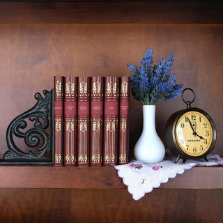 Jane Austen book set displayed on a wooden shelf alongside a black ornate iron book end, a white vase with lavender flowers, a decorative clock, and an embroidered floral cloth. The deep red covers with ornate gold detailing stand elegantly at the center, blending classic charm with timeless decor.