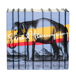 A set of eight blue hardcover books by Ernest Hemingway arranged side by side to form a continuous cover illustration. The cover features an elephant standing against a background with horizontal yellow and red stripes, with Ernest Hemingway's signature in white spanning across the spines. Each spine displays the title of a book.