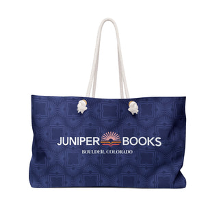 Large canvas blue beach bag with white rope handles. Bag fabric is dark blue with lighter Juniper Books logomark pattern overlay. Juniper Books company name, location, and logo across the side in white.