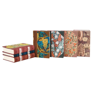 A collection of six beautifully bound books with decorative covers featuring intricate floral and nature-inspired patterns. Three books are stacked horizontally on the left, while four stand upright on the right. The spines of the horizontal books have a classic design with labels and ornamental elements. The upright books have colorful, artistic covers with detailed botanical motifs, some with faux leather edges.