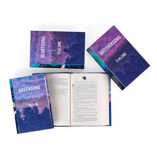 A top view of four hardcover books from the Green Creek series by TJ Klune, arranged on a white surface. The covers feature a misty forest landscape with a purple and blue color scheme, with titles including Heartsong, Ravensong, and Brothersong visible. One book is open, showing the interior pages with text.