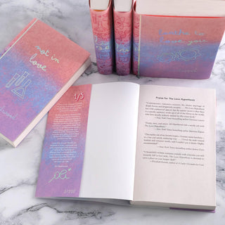 A set of five Ali Hazelwood novels with custom designed book jackets laid on a white marble surface. One book in the center is open revealing the inner front cover of Book One, titled "The Love Hypothesis." The inner flap of the book jacket includes is the author's signature in the same holographic foiling as the front cover, and the number 1/100 to showcase the edition number of this limited edition custom book jacket set.