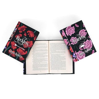 A flat lay photograph of three books by Lauren Roberts on a white background. The center book is open, revealing printed text, while the other two are closed and placed diagonally on either side. The book on the left, titled Reckless, features a black cover adorned with red flowers and elegant white typography. The book on the right, titled Fearless, has a similar black cover design but with pink roses and a white crown illustration.