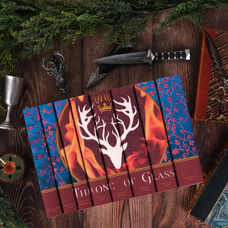 This image highlights the Throne of Glass custom book set displayed in a flat-lay composition. The books are arranged on a wooden surface surrounded by thematic props, including a dagger, a stag ornament, a metal goblet, and greenery, evoking a medieval and fantasy atmosphere. The spines feature the vivid antlered crown illustration and fiery design, complemented by the intricate blue floral patterns, adding to the rich and immersive aesthetic of the series.