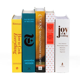 A row of five classic cookbooks standing upright without their dust jackets, revealing their hardcover bindings. The books include 'How to Cook Everything' by Mark Bittman in a bright yellow cover, 'The Recipes of Record' by Amanda Hesser in a blue cover with a black emblem, 'Cook’s Revolutionary Recipes' by America’s Test Kitchen in a gold and brown cover, 'Mastering the Art of French Cooking' by Julia Child in a white cover with red and blue accents, and 'Joy of Cooking'