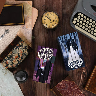 A vintage-themed setup featuring Nancy Drew and Hardy Boys book sets with custom illustrated covers. The Nancy Drew books are stacked to form an image of a silhouetted figure holding a magnifying glass against a purple background, while the Hardy Boys books form an image of two silhouetted figures walking through a forest against a blue background. The scene includes props like an antique clock, an old-fashioned typewriter, marbled and leather-bound books, handwritten letters, and maps, on a wood background