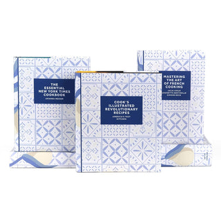 A set of three cookbooks with decorative slipcases featuring a blue and white tile pattern. The titles displayed are 'The Essential New York Times Cookbook' by Amanda Hesser, 'Cook’s Illustrated Revolutionary Recipes' by America’s Test Kitchen, and 'Mastering the Art of French Cooking' by Julia Child, Louisette Bertholle, and Simone Beck. The books are arranged in a staggered manner, with one in the foreground and two slightly behind.