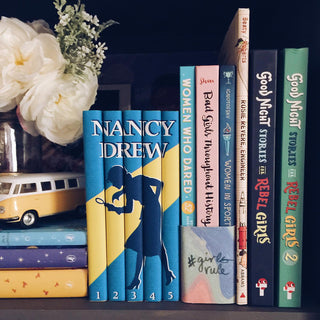 Install shot of the blue and yellow vintage Nancy Drew set from Juniper books on a shelf surrounded by white flowers, and other stories that empower and inspire young girls. 