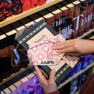 Person cleaning an Outlander Botanical jacketed book with a patterned cleaning cloth with Juniper Books branding in front of shelves with jacketed books.