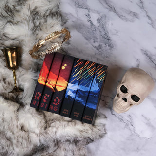 This image shows a flat-lay arrangement of all the books in the Red Rising Series by Pierce Brown placed on a fur rug against a marble background. The book spines display a vibrant, cohesive illustration: warm red and orange hues on the left depict a Martian landscape with two suns, while cool blue tones and streaking meteors appear on the right. A golden laurel crown rests above the books, and a gold chalice is set to the left. On the right side of the image, a human skull with hollow black eye sockets.