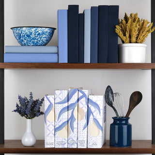 A stylish kitchen shelf display featuring a coordinated set of five cookbooks with blue and white dust jackets showcasing a seamless design of leafy branches and pears. The books are arranged alongside a deep blue jar holding kitchen utensils, including a whisk and wooden spoons. Above, another shelf holds blue-toned books, a blue patterned ceramic bowl, and a vase filled with golden dried flowers. A small white vase with lavender sits next to the cookbook set.