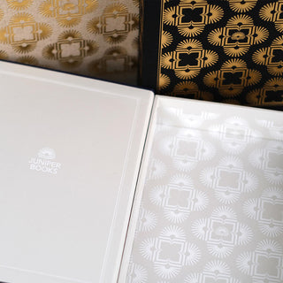 Close up of the interior of the dust jacket storage boxes. The inside is pale grey and the inner box features the same symmetrical pattern in white. The inner lid of the box has the Juniper Book's logo centered in white.