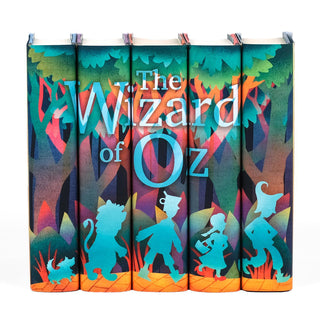 A set of five hardcover books with book jackets arranged side by side to form a continuous illustration of 'The Wizard of Oz.' The spines feature silhouettes of iconic characters: Toto, the Cowardly Lion, the Tin Man, Dorothy, and the Scarecrow, walking along a yellow brick road through a vibrant, stylized forest. The title 'The Wizard of Oz' appears in large, bold letters across the spines, blending with the forest background in shades of teal, orange, and purple.