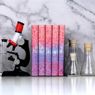 A set of five Ali Hazelwood novels in custom designed book jackets on a white surface with a white marble backdrop. The books are displayed among beakers and a microscope. A continuous design is visible across the spines of all five books, with the name 'Ali Hazelwood' written in a playful lowercase cursive font across all five books. The cursive lettering stands out with a shimmering, elegant foiling effect. The design features a pink, blue, and purple gradient peppered with scientific and flirty symbols.