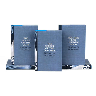 Three Hardy Boys books by Franklin W. Dixon displayed upright, each with a blue textured cover and white text. The titles are 'The House on the Cliff,' 'The Secret of the Old Mill,' and 'Hunting for Hidden Gold.' Two books are placed horizontally in the background, partially visible, with edges showing a coordinated design that continues onto the spines.