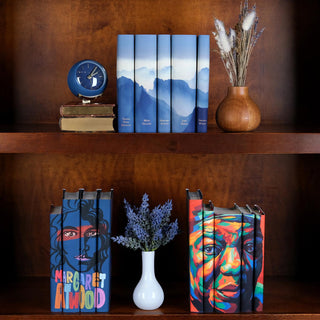 A wooden bookshelf is neatly arranged with decorative book sets and accessories. On the top shelf, a collection of five hardcover books with blue-toned mountain landscape dust jackets is displayed upright, alongside a small stack of vintage books with a blue clock on top and a wooden vase holding dried flowers. The bottom shelf showcases two sets of books with vibrant, artistic dust jackets One set features a bold, colorful portrait of Margaret Atwood and the other a colorful portrait of Maya Angelou.