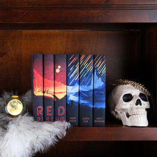 A bookshelf decorated with all 6 books in the Red Rising series by Pierce Brown. The book spines form a cohesive design, with vibrant, contrasting colors—on the left side, warm red and orange hues depict a Martian landscape beneath a two-sun sky, and on the right side, cool blue and green tones show a Mecurian City an iron rain. Beside the books is a white skull wearing a golden laurel, a faux fur pelt drapes over the shelf, and a glowing moon sits to the left of the books.