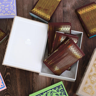 A collection of Jane Austen hardcover books with ornate gold foil jackets displayed on a wooden table. A neutral box with and white interior and 'Juniper Books' branding is open in the center, containing a couple of the Jane Austen dust jackets with deep red covers and intricate gold foil designs. Other books in the set are scattered around the box, showcasing their bright covers.