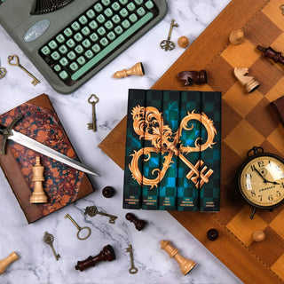 The Inheritance Games Series by Jennifer Lynn Barnes from Juniper Book sits on a wooden chess board sitting on white marble background. The set is surrounded by wooden chess pieces, ornate gold keys, antique leather books, a dagger, an antique clock, and a typewriter.