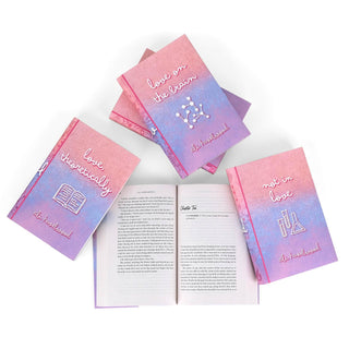 A set of five Ali Hazelwood novels in custom designed book jackets featuring a gradient of pink, blue, and purple photographed in a casual display from above on a white background. One book at the center is opened to reveal the text within, and is surrounded by the other books from the set. The titles of three covers are visible, featuring playful lowercase cursive. Below each title, there are various STEM-related iconography illustrated in clean white lines.