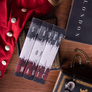 Flat lay of Shades of Magic set from Juniper Books. Set sits on antique leather books surrounded by a red coat, book on London, and a pocket watch set against wood background.