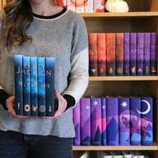 Person holding books titled 'Percy Jackson and the Olympians' in front of a bookshelf with similar books.