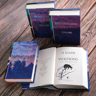 A collection of four hardcover books from the Green Creek series by TJ Klune is displayed on a wooden surface. Three books, 'Heartsong,' 'Brothersong,' and 'Ravensong,' are standing upright or laid flat, each with a cover featuring a forest silhouette under a sky with blue and purple hues. The fourth book, 'Wolfsong,' is open to show the signed title page with an illustration of a wolf and the author’s signature in black ink.