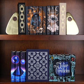 A wooden bookshelf displaying multiple sets of books with artistic dust jacket covers by Juniper Books. On the top shelf, two decorative bookends frame three sets: a black and beige dust jacket box on each end, a set with a skull and foliage design, and a set with a large eye illustration. The bottom shelf features three sets: a cosmic pyramid design, a navy blue dust jacket box with a silver pattern, and a set titled 'The Lunar Chronicles' with a circuit board design.