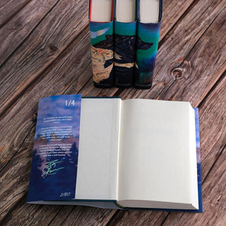 An open book displayed on a wooden surface, with the dust jacket flap showing on the left side. The flap contains a quote, a message, and TJ Klune's signature in holographic foil. Behind the open book are three hardcover books with illustrated dust jackets featuring wolves and nature scenes that align to form a continuous image across the spines.