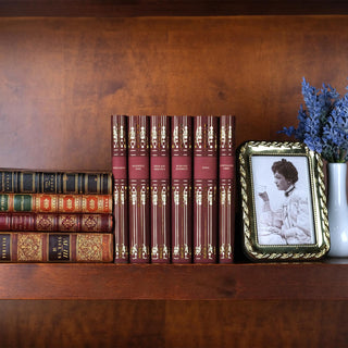 Jane Austen book set displayed on a wooden shelf alongside antique leather books, a photo of a woman drinking tea in a gold picture frame, and a white vase with lavender flowers. The deep red covers with ornate gold detailing stand elegantly at the center, blending classic charm with timeless decor.