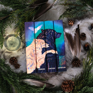 A beautifully arranged display of four books with illustrated spines that form a continuous image of a light-colored wolf embracing a dark wolf, set against a gradient blue-green background. The book titles are at the base of each spine in holographic foil. The scene is surrounded by natural elements, including pine branches, pinecones, large feathers, and a moon-like glass orb, all placed on a soft, fur-like fabric.