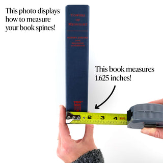 A demonstration of how to measure the spine of a book using a tape measure. The book shown is Towers of Midnight by Robert Jordan and Brandon Sanderson, with a navy spine featuring red text. A tape measure held along the spine shows the width as 1.625 inches. Text on the image reads, "This photo displays how to measure your book spines!" and "This book measures 1.625 inches!