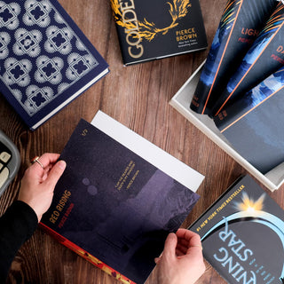 Two hands wrapping Red Rising in artistic Juniper Books dust jackets on a wooden background. Surrounded by Golden Son, Morning Star, a navy and silver Dust Jacket Storage Box, and an open storage box holding Pierce Brown Red Rising Extension dust jackets.