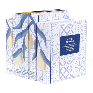 A side-angled view of a coordinated set of five cookbooks with matching dust jackets featuring a seamless design of blue leafy branches and stylized pears on a blue and white tiled background. The front cover of Joy of Cooking is visible, displaying a blue title box with white text listing the authors: Irma S. Rombauer, Marion Rombauer Becker, Ethan Becker, John Becker, and Megan Scott.