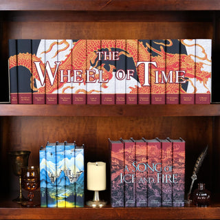 The Wheel of Time Book Set sitting on a dark wood shelf above a Tolkien's Epic Journey and A Song of Ice and Fire Book Set from Juniper Books. The book sets are surrounded by goblets, a candle, and a quill and ink pot. 