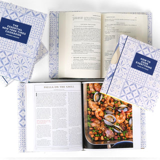 An assortment of open and closed cookbooks with decorative book covers featuring a blue and white tile pattern. The books shown include 'The Essential New York Times Cookbook' by Amanda Hesser and 'How to Cook Everything' by Mark Bittman. One open book displays a recipe for 'Paella on the Grill' alongside a photograph of a seafood and rice dish with shrimp, clams, peas, and other ingredients. Another open book reveals a recipe for 'Chicken Fricassee.'